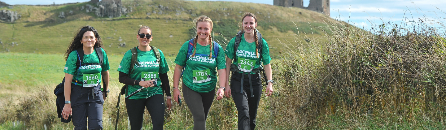 A group of four hikers are walking along a grassy path. Behind them are grassy fields and old ruins.